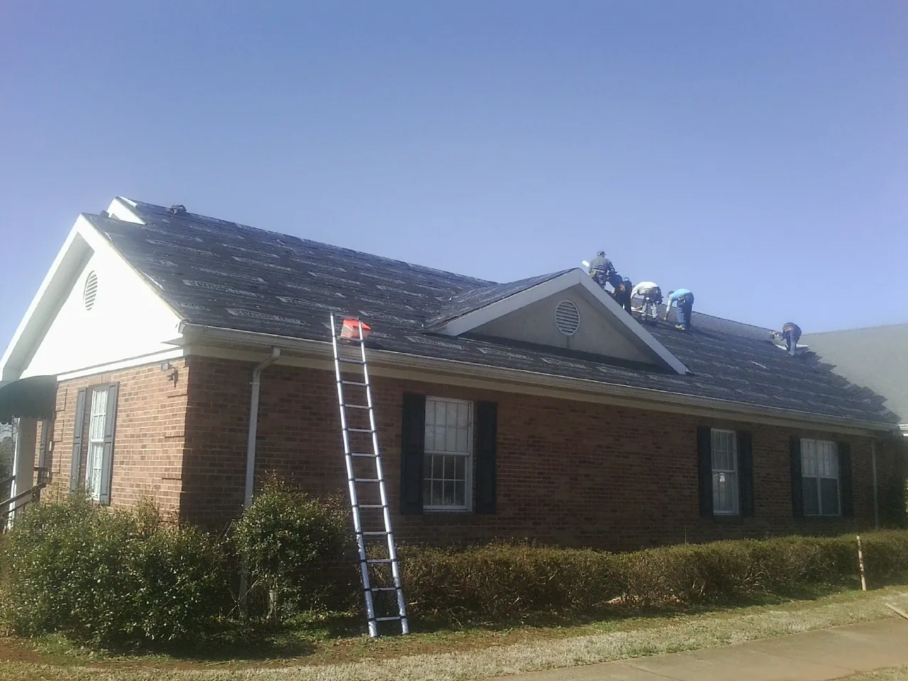 Crew tearing shingles off a brick ranch roof with a ladder set — Emergency Roof Repair work by our crew in Midfield