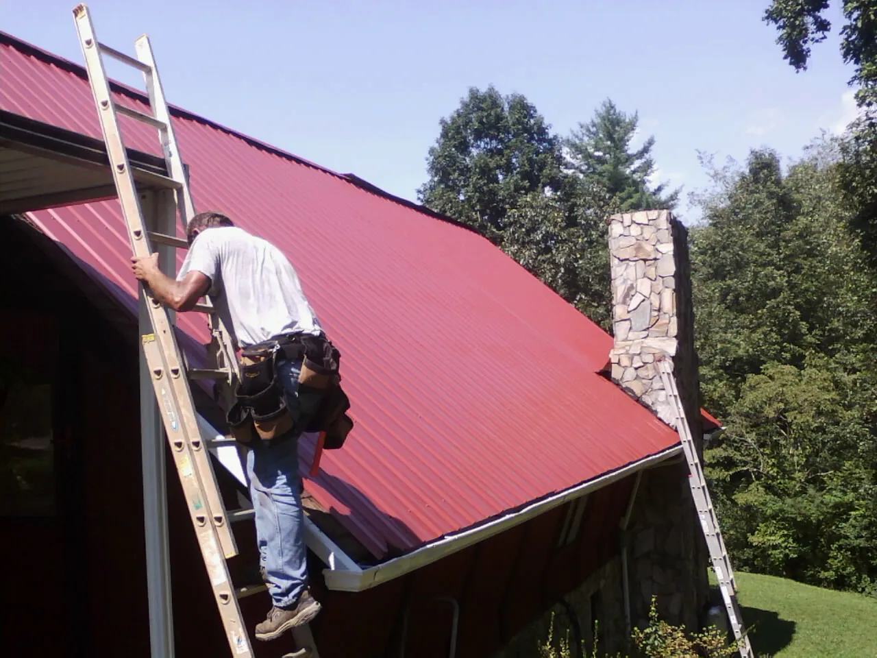 Roofer on a ladder beside a red metal roof and stone chimney — Soffit & Fascia Repair by Roofing Brothers in Midfield